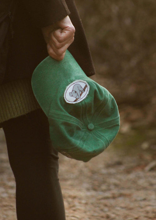 Close up of a hand holding the brim of a green baseball cap with a vintage style patch of a koala illustration hanging off a branch with frayed edges