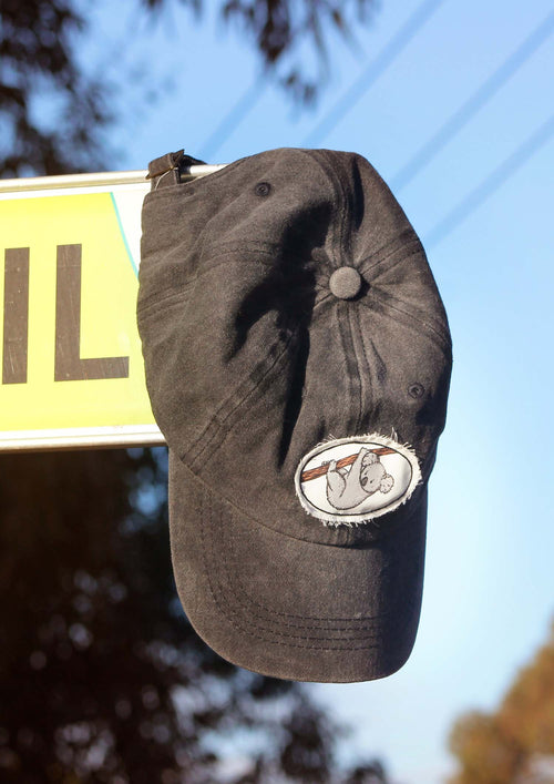 Black baseball cap hanging of a street sign in Raymond Island. The hat has a frayed edge patch with an original illustration of a koala hanging from a branch.